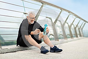 Mature sportsman having break, drinking water, using smartphone