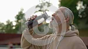 Mature sporty man drinking water from the sport bottle while walking. Elderly male feeling thirsty. Aging youthfully