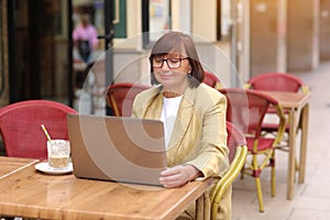 Mature stylish businesswoman in eyeglasses works on laptop while sitting with a coffee drink at European cafe outdoors