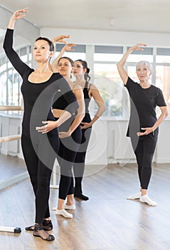 Mature teacher conducts class with girls in ballet studio, observes doing forth position exercise.