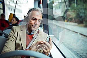 Mature tired businessman with heaphones and smartphone travelling by bus in city.