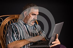 Mature Ukrainian peasant sitting in wicker chair and typing on a laptop PC