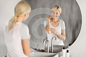 Mature Woman Applying Cream On Elbow While Standing Near Mirror In Bathroom
