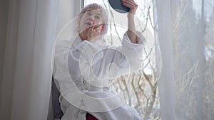 Mature woman applying facial moisturizer sitting on windowsill at home. Portrait of confident Caucasian lady taking care