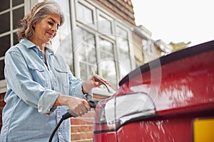 Mature Woman Attaching Charging Cable To Environmentally Friendly Zero Emission Electric Car At Home