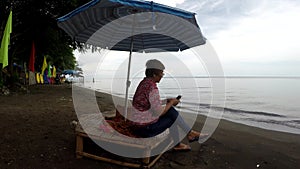 Mature woman browses internet with the use of mobile smartphone while sitting cross legged under parasol sunshade
