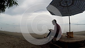 Mature woman browses internet with the use of mobile smartphone while sitting cross legged under parasol sunshade