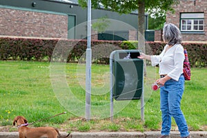 Mature woman in casual clothing depositing her dog`s waste in a public rubbish bin on the street