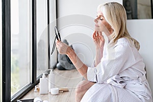 Mature woman checking result after skincare procedures, looking at hand mirror