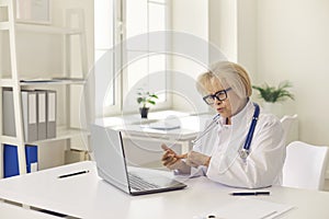 Mature woman doctor in uniform and glasses consulting patient online on laptop