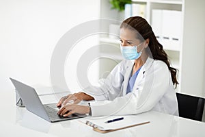 Mature woman doctor wearing face mask typing on laptop keyboard