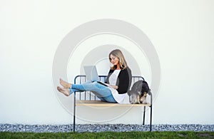 Mature woman with dog working in home office outdoors on bench, using laptop.