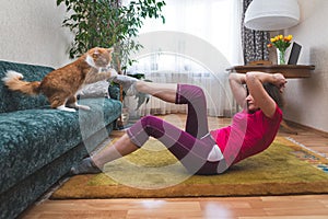 Mature woman doing abdominal exercise, her ginger cat plays with the owner`s leg