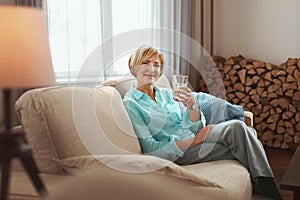 Mature Woman Drink. Middle Age Female In Casual Clothes Has Fresh Water With Lemon Juice On Couch At Home.