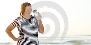 mature woman drinking water from bottle after fitness exercises or yoga on beach by sea. fitness and healthy lifestyle
