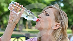 Mature woman drinking water from bottle in park, maintaining water balance