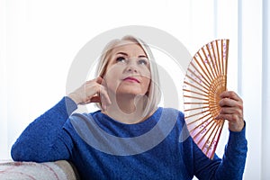 Mature woman experiencing hot flush from menopause. A woman in her fifties sitinds fanning herself with a handheld fan, her face photo