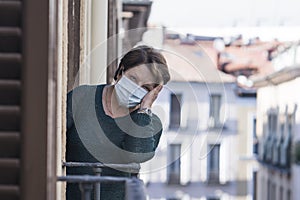 Mature woman with face mask sad and scared at home balcony during covid19 pandemic lockdown looking to the street confused and