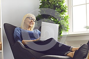 Mature woman with glasses sitting in an easy chair and working at home using a laptop.
