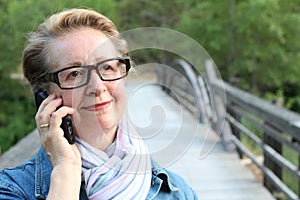 Mature woman with glasses waiting your call. Dressed in a denim jacket, a senior Caucasian lady is standing by river bridge