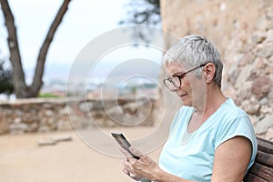 Mature woman with gray hair and glasses looks at her smartphone sitting in a park in summer