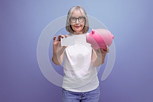 mature woman with gray hair keeps her savings on a credit card and in a piggy bank on a studio background with copy