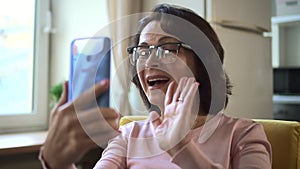 Mature woman has online talk and uses smartphone, sits in home room indoors.