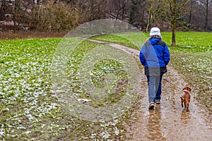 Mature woman with her dog walking a wet dirt trail between a valley with green grass, her back to the camera