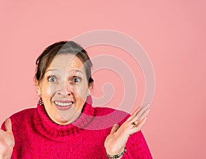 A mature woman in her early fifties with a pink turtleneck looking at the camera exasperated with hands in the air