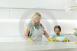 Mature woman and her son kid in protective gloves are smiling, using detergent and dusters while cleaning furniture at home