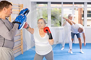 Mature woman with her trainer at gym. Woman wearing boxing gloves exercise and punch to pads for boxing