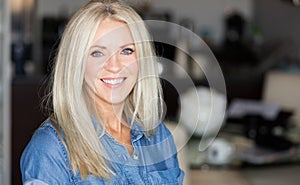 Mature woman in a jeans shirt smiling at the camera. She is in her kitchen relaxing