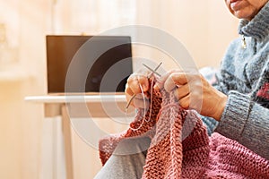 Mature woman knits a blanket and watching online lessons in a laptop.