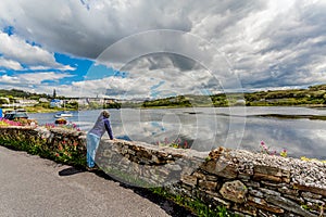 Mature woman leaning on a stone fence looking at the pier in the port of Clifden with anchored boats