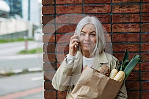 Mature woman making phone call, going home from supermaket with gorceries. Beautiful older woman with gray hair standing