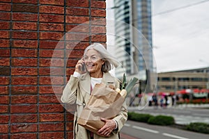 Mature woman making phone call, going home from supermaket with gorceries. Beautiful older woman with gray hair standing