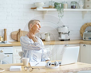 Mature woman massaging her neck, feeling tired while working or studying on laptop online at home