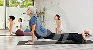 Mature woman performing Bhujangasana or Cobra pose during yoga class with female group in fitness studio