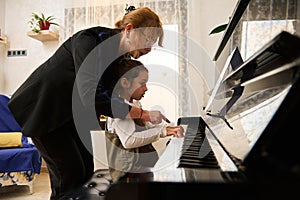 Mature woman pianist teacher giving piano lesson to a little school girl, teaching the position of fingers on piano keys