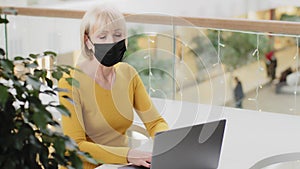 Mature woman professional worker freelancer in protective mask sitting in workplace typing on laptop modern middle aged