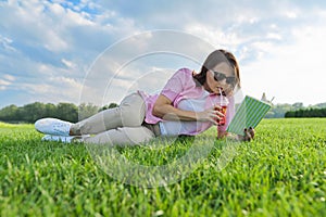 Mature woman reading book, female lying on green grass with drink