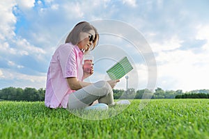 Mature woman reading book, female sitting on green grass with drink