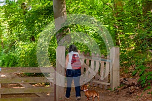 Mature woman with a red backpack and her dog opening the wooden gate to enter the forest with abundant vegetation