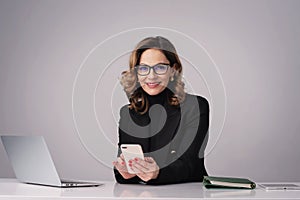 Mature woman sitting at desk and using laptop and smartphone against isolated background