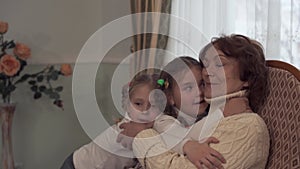 Mature woman sitting in a rocking chair and her small cute granddaughters came to her to kiss and gently hug