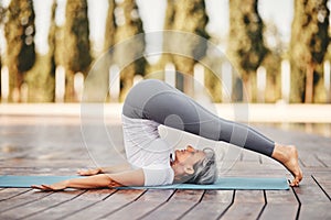 Mature woman in sports wear practicing yoga Halasana on mat outdoors outdoors photo