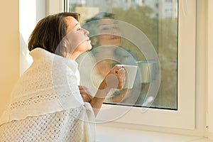 Mature woman standing near the window with cup of hot drink under warm knitted woolen blanket looks dreamily out the window,
