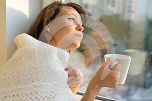 Mature woman standing near the window with cup of hot drink under warm knitted woolen blanket looks dreamily out the window,