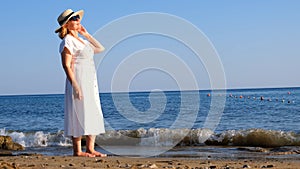 mature woman in a straw hat and white dress enjoying the sun walking along the blue sea coast on a sunny summer day