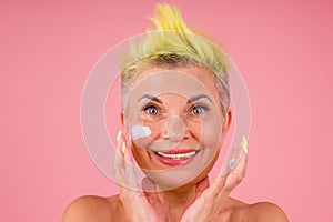 Mature woman with stylish yellow dyed hair and bare shoulders applying anti-aging cream on pink studio background